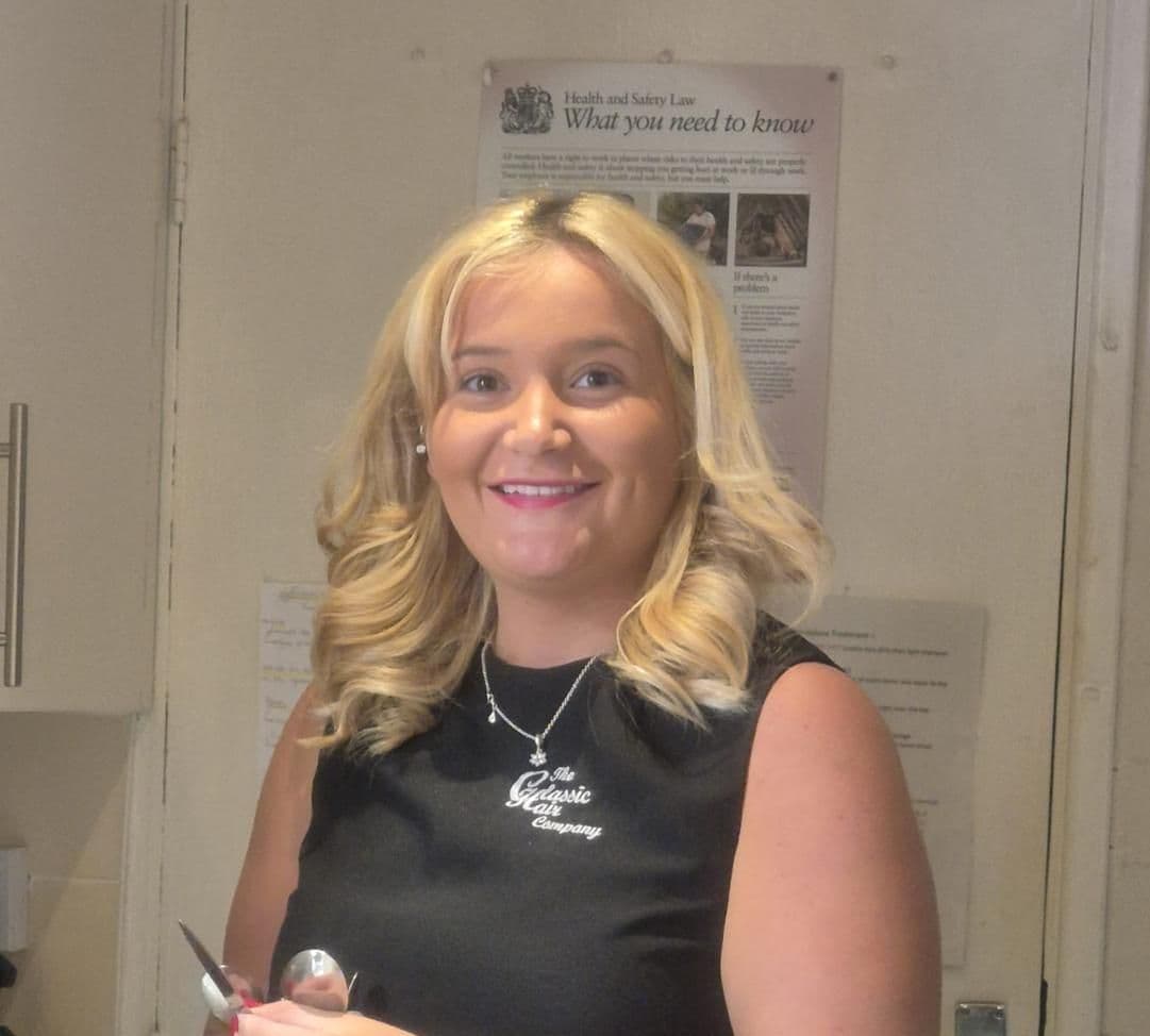 Tillie Mynett, Stylist at The Classic Hair Company in Tilbury, Essex, smiling in the salon wearing her uniform.